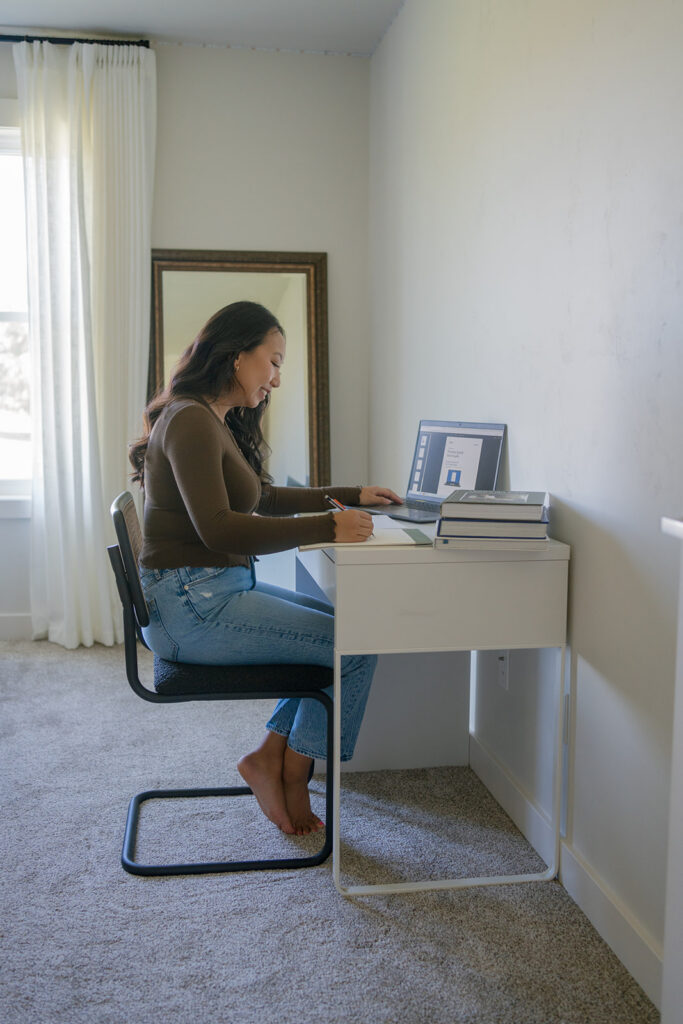 A student writes at a desk by a window, with a laptop and books in front of her.