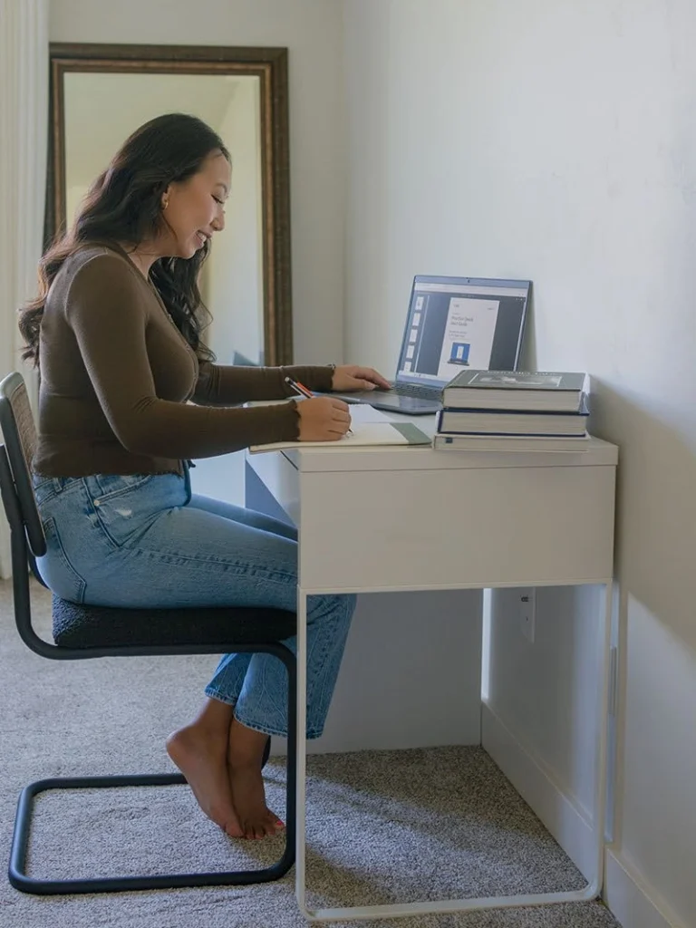 A student writes and uses a laptop at her desk.